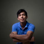Young Hispanic man smiling confidently in a blue shirt against a dark background.